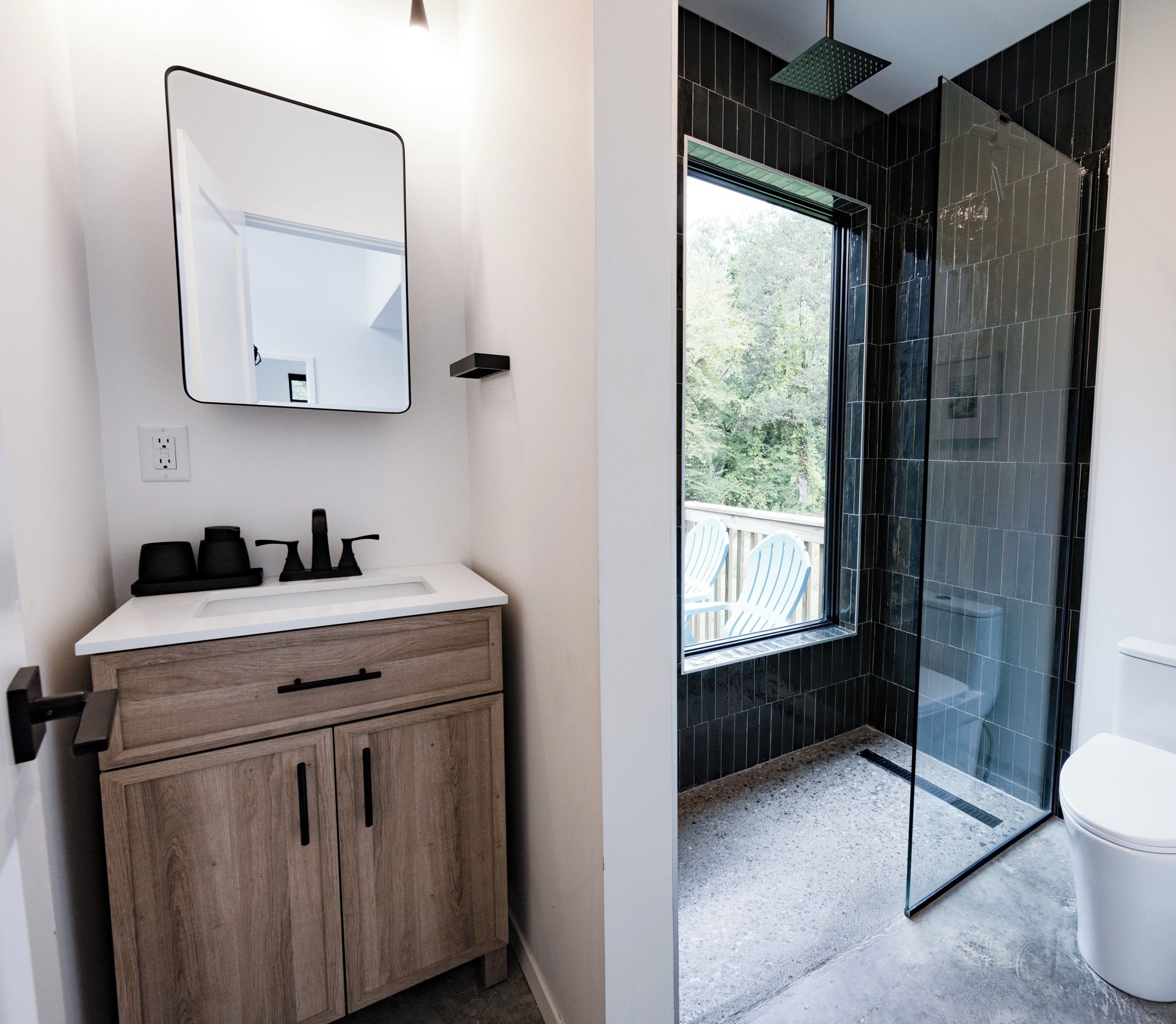 A wood vanity, matte black fixtures, and a walk-in rain shower wrapped in deep green glazed tile. A tall window looks out to the trees, so every shower feels a little like showering outdoors.