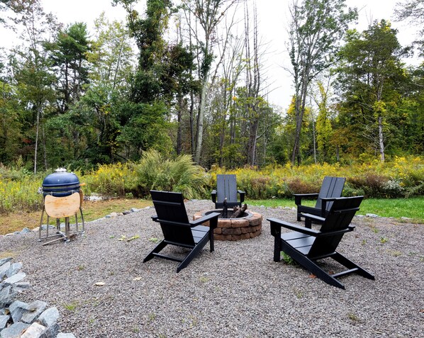 Un'area relax esterna con barbecue, sedie e un braciere, circondata da ghiaia e alberi.
