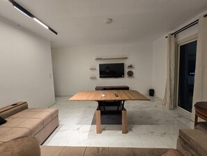 A modern living room with a wooden dining table, a sofa, a flat-screen TV mounted on the wall, and a sliding glass door leading to an outdoor area. - Hurghada Lavanda Beach Resort (Hurghada)