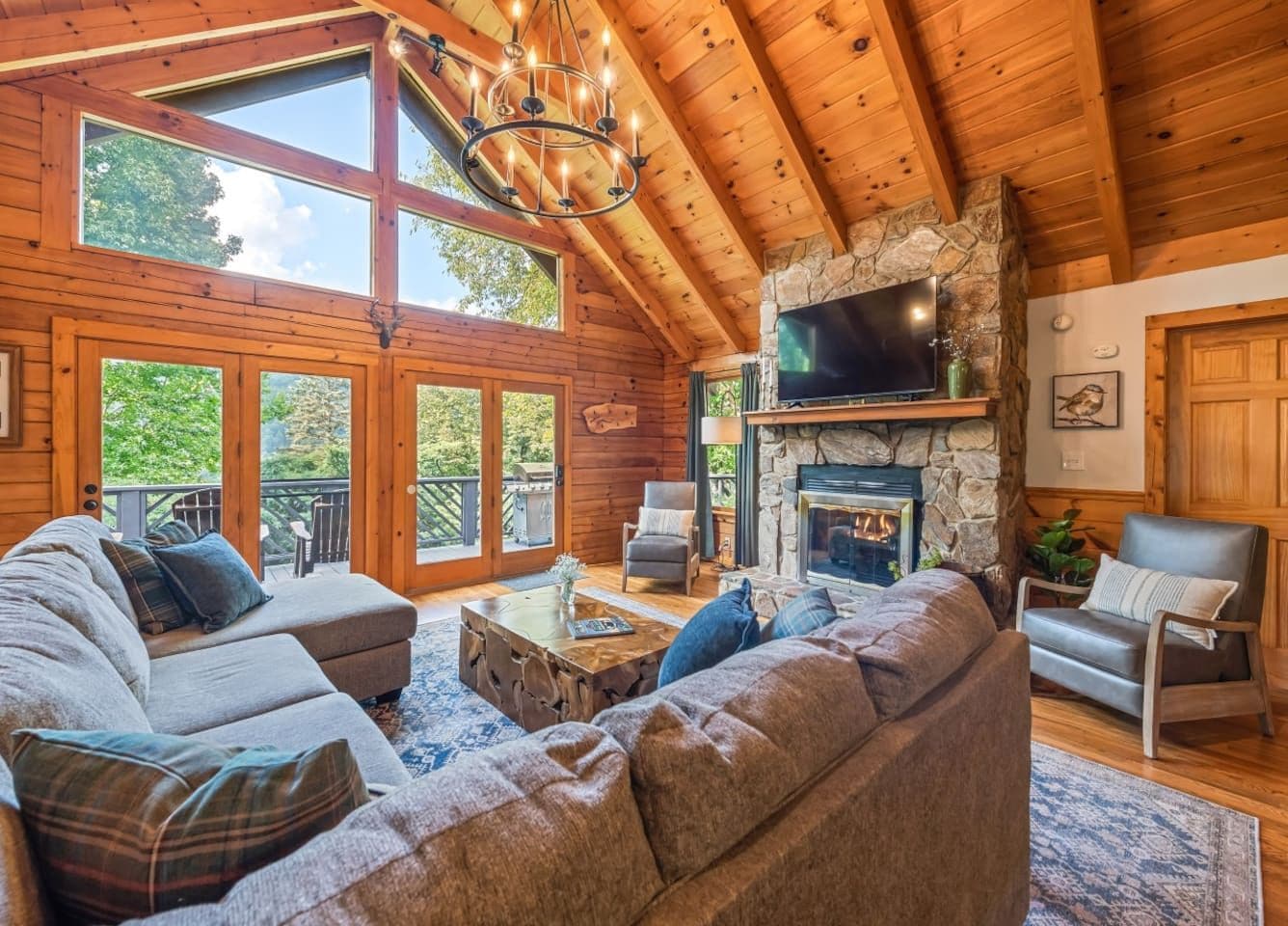 A log cabin interior with a stone fireplace, a large sectional sofa, and a television mounted on the wall.