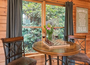 A wooden cabin interior with a chessboard on a round table, two chairs, a vase with flowers, and a window with curtains. - Rocky Top Retreat-Hot Tub, Game Room, Garage, View (Maggie Valley)