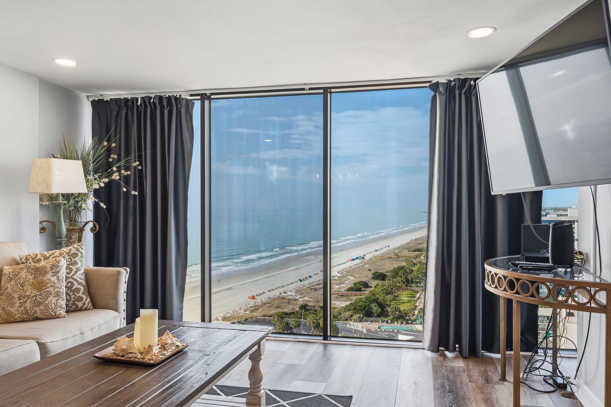 A living room with a large window overlooking a beach, a flat-screen TV, and a coffee table with decorative items.