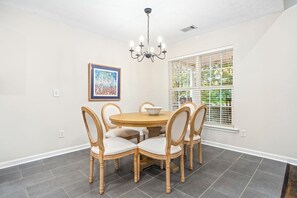 Een eetkamer met een ronde tafel, zes stoelen, een kroonluchter, een lijst met een afbeelding en een raam met jaloezieën.