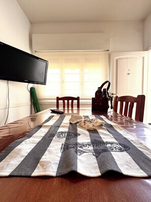 A dining area with a wooden table, chairs, a television mounted on the wall, and a window with blinds.