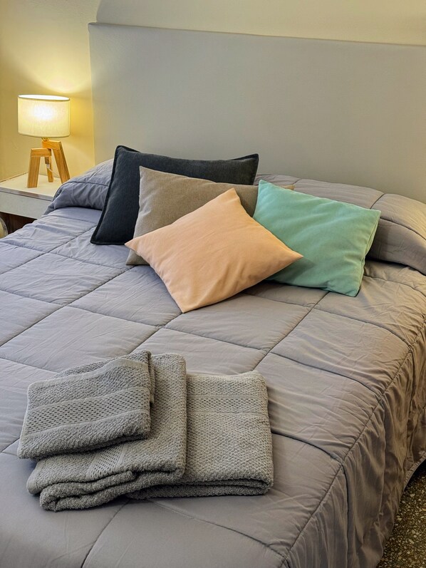 Un lit avec une couette grise, plusieurs oreillers de différentes couleurs et une couverture pliée.