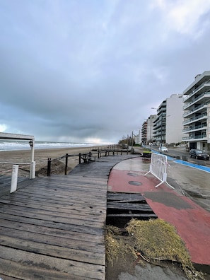 A wooden boardwalk leading to a beach, with a red path and a barrier on the right.