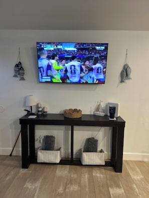 A television mounted on a wall displaying a sports event, a dark wooden console table with decorative items, and two storage baskets below. - Chasing Sunsets
Cozy 1-bedroom  2 bath  with 2 beds and All new furnishings. (Galveston)