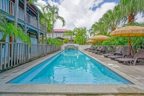 Una piscina con sillones de descanso y sombrillas, rodeada de palmeras y un edificio con techo rojo.