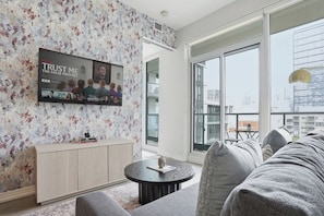 A modern living room with a large TV mounted on a floral wallpaper wall, a sofa, and a round coffee table. - City View Condo, Walk to Eaton Centre, Sleeps 6! | Parking Available (Toronto)
