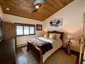A bedroom with a wooden ceiling, a bed with a patterned bedspread, a nightstand with a lamp, and a window with a view of the outdoors. - New Listing! 3BR Luxury Big Sky | Mountain Retreat (Big Sky)