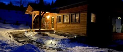 A wooden cabin with a covered porch and a snow-covered landscape at dusk.