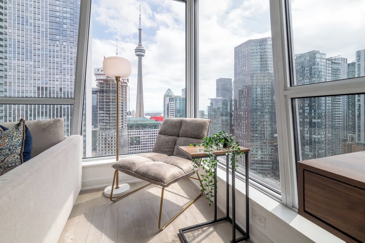 A modern living room with a sofa, a small table with a plant, and a lamp. A cityscape with tall buildings and a tower is visible through large windows.