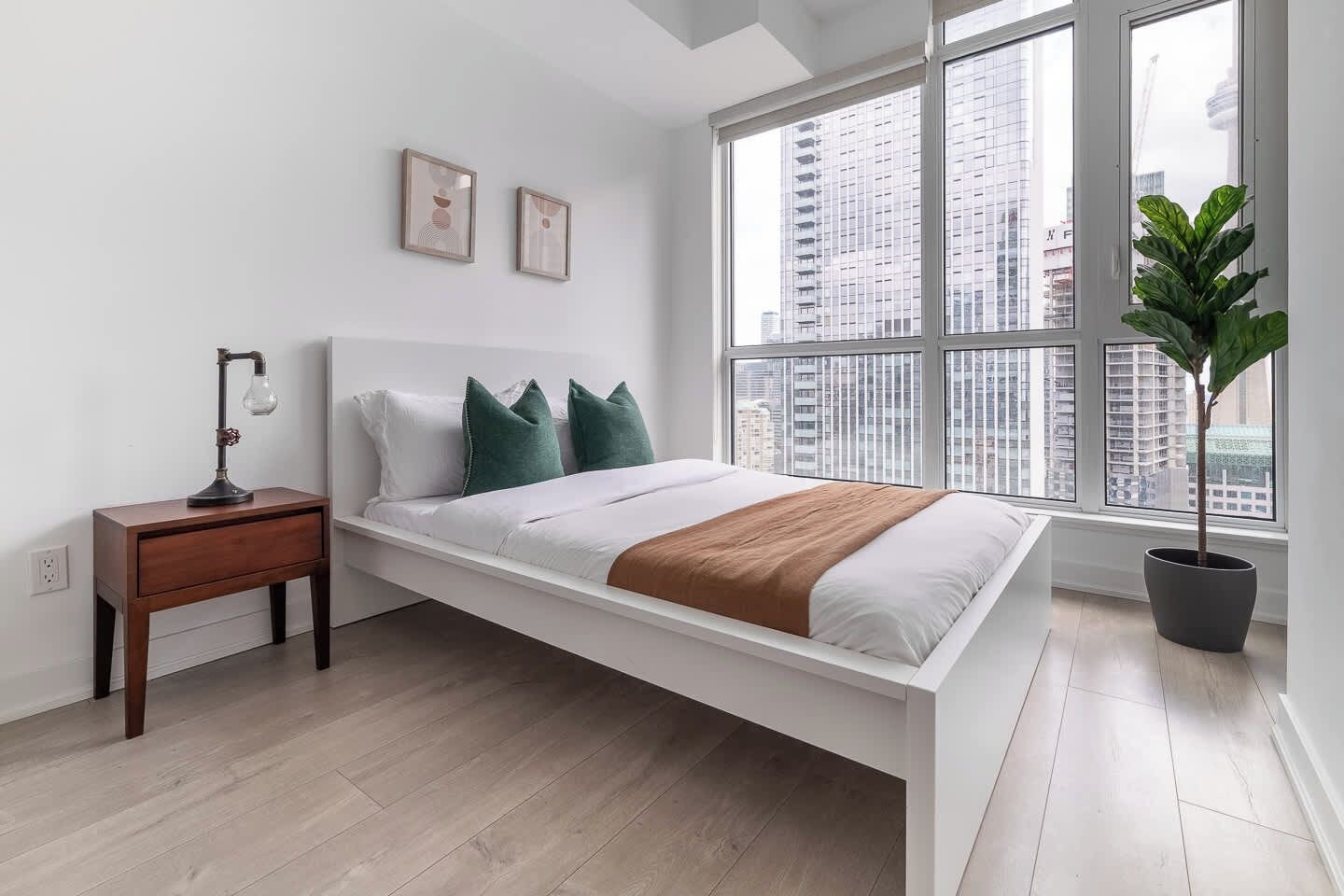A modern bedroom with a bed, a nightstand, a lamp, a potted plant, and a large window with a city view.