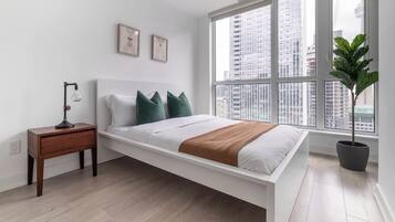 A modern bedroom with a bed, a nightstand, a lamp, a potted plant, and a large window with a city view.