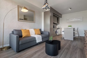 A modern living room with a grey sofa, a black coffee table with a plant, a large mirror, and a kitchen area in the background. - Modern Cozy Condo in the Heart of Toronto! | Parking Available (Toronto)