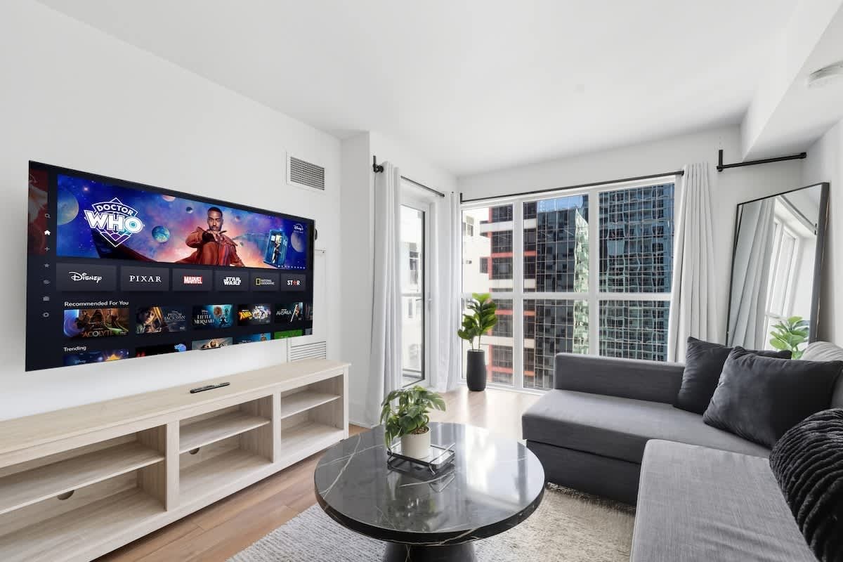A modern living room with a flat-screen TV displaying a streaming service interface, a grey sofa, a coffee table, and a large window offering a view of the city.