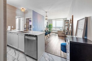 A modern kitchen with a dishwasher, a dining area with a blue chair, and a large window offering a city view. - Luxe Downtown 1BR Suite, Workspace & 55" TV (Toronto)