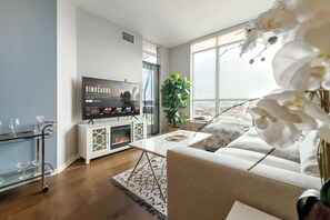 A modern living room with a flat-screen TV, a fireplace, a sofa, and a large window offering a city view. - Luxe Downtown 1BR Suite, Workspace & 55" TV (Toronto)