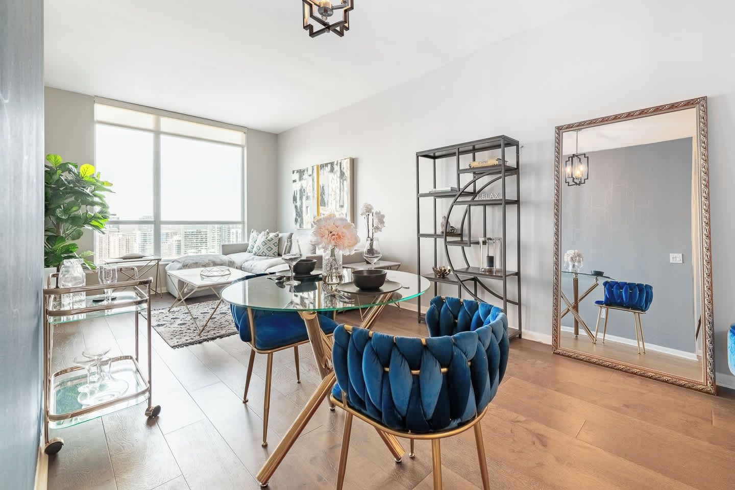 Un salon moderne avec une table à manger en verre, des chaises rembourrées en tissu bleu, une bibliothèque et un grand miroir.