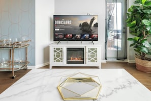 A living room with a fireplace, a television displaying 'The Dinosaurs', and a glass coffee table. - Luxe Downtown 1BR Suite, Workspace & 55" TV (Toronto)