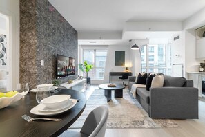 Ein modernes Wohnzimmer mit einem grauen Sofa, einem Esstisch mit Tellern und Besteck, einem flachbildfernseher an einer strukturierten Wand und einem großen Fenster, das für natürliches Licht sorgt.