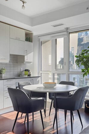 A modern kitchen with white cabinets, a round dining table with four chairs, and a view of the city through large windows. - Stylish TO Condo CN Tower View Walk Score 99 | Parking Available (Toronto)
