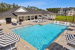 A swimming pool with lounge chairs and umbrellas, a covered patio area, and a building with a gabled roof. - Wildlight Retreat · Pool · Pickleball · Canopy Bed (Yulee)