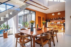 A modern dining area with a wooden table and chairs, a vase with flowers, and a view of a pool outside. - Green Roof Villa Pandawa (KAB. BADUNG)