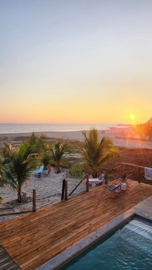 Une terrasse en bois avec des chaises longues et une piscine, offrant une vue sur la plage au coucher du soleil.