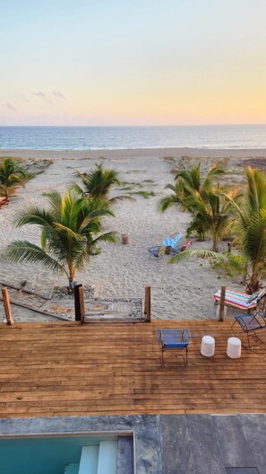 A wooden deck overlooking a beach with palm trees, a pool, and lounge chairs.