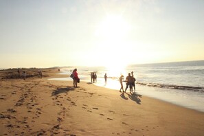 Uma praia com pegadas indo em direção à água, pessoas caminhando e o sol se pondo ao fundo.