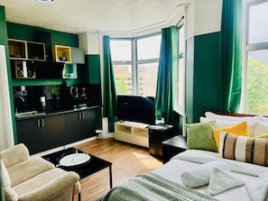 Habitación de hotel compacta con kitchenette, una cama con varias almohadas, sofá y ventana con cortinas.