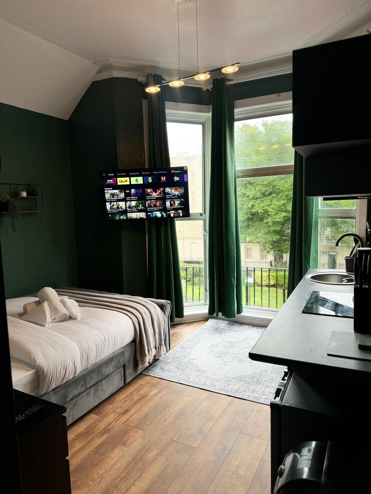 A hotel room with a bed, a TV mounted on the wall, a window with curtains, and a kitchenette area.