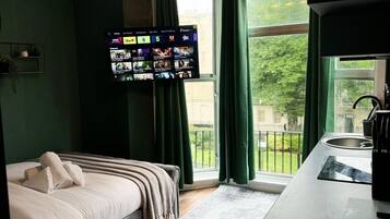 A hotel room with a bed, a TV mounted on the wall, a window with curtains, and a kitchenette area.