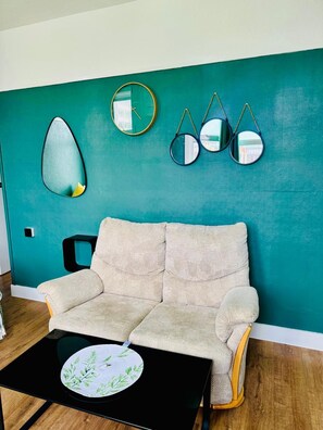 A teal-colored wall with a round mirror, a clock, and three hanging mirrors. A beige sofa is positioned against the wall, and a black coffee table with a leaf-patterned coaster is in front of it. - Dudy Studios at Brighton city center (Brighton)