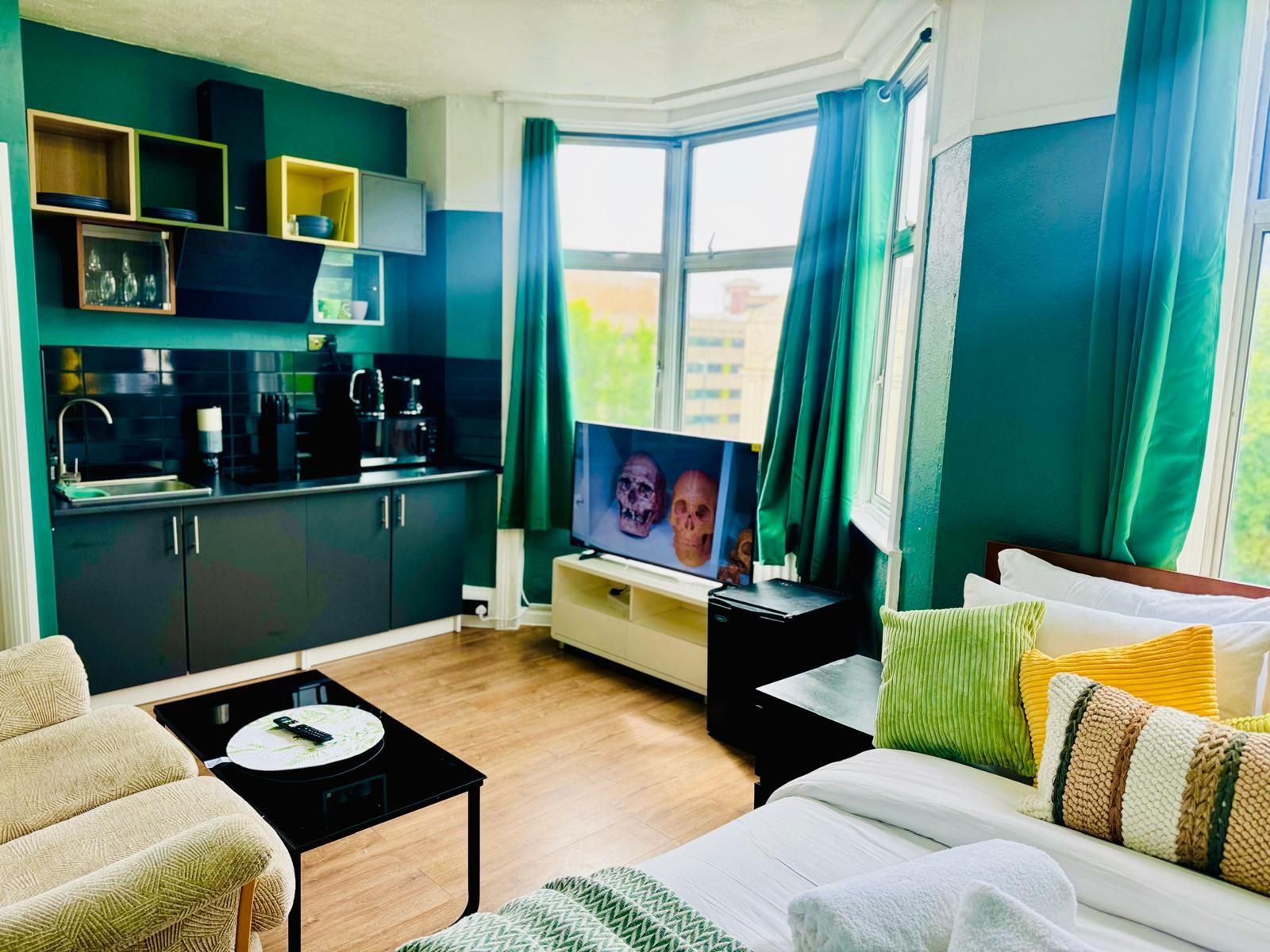 A modern living room with a sofa, a coffee table, a TV, and a kitchen area with cabinets and a coffee machine.