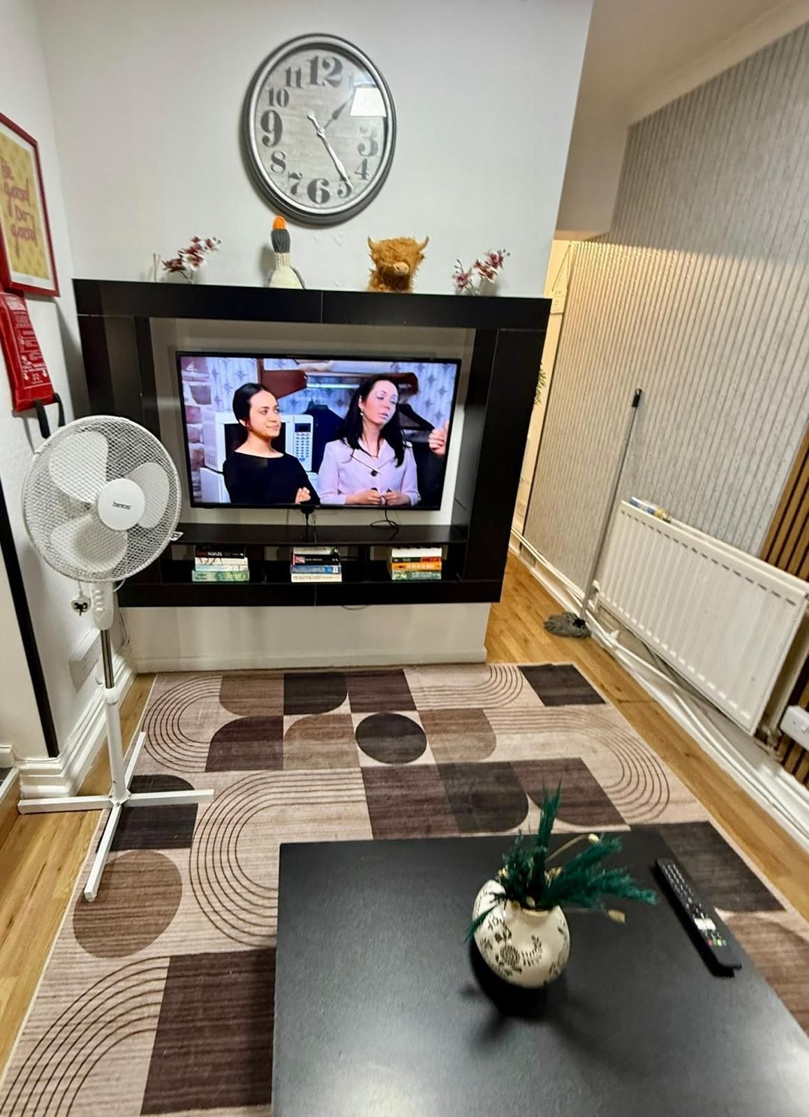 A living room with a flat-screen TV, a clock, a fan, a coffee table, and a radiator.