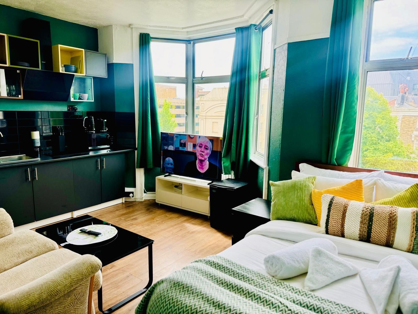 A modern hotel room with a kitchenette, a television, a bed with multiple pillows, and a view of the city through large windows.