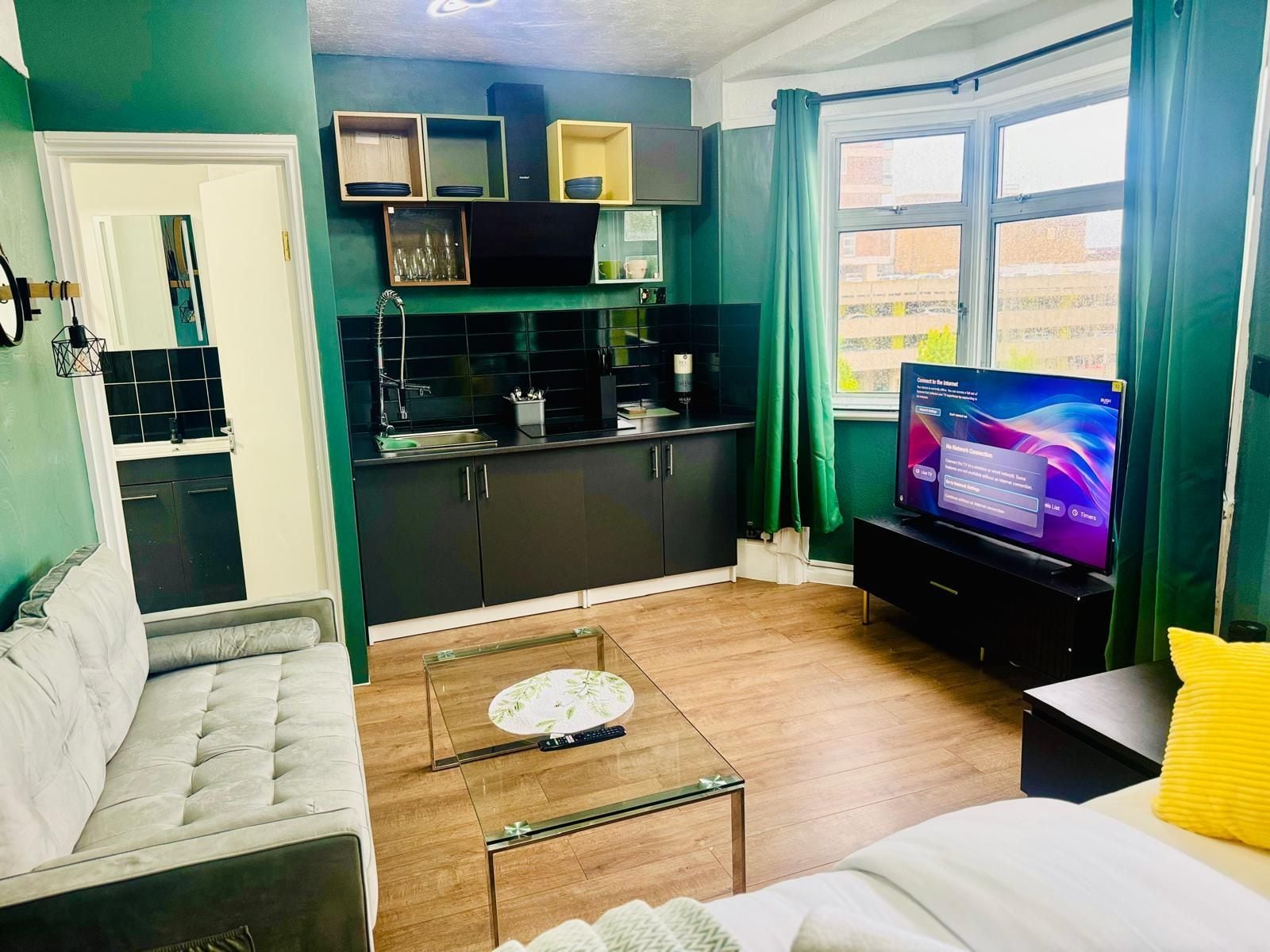 A living room with a sofa, a coffee table, and a television. There is a kitchen area with cabinets and a window.