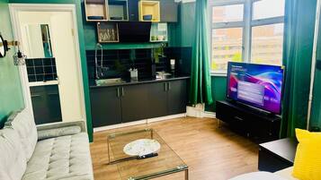 A living room with a sofa, a coffee table, and a television. There is a kitchen area with cabinets and a window.
