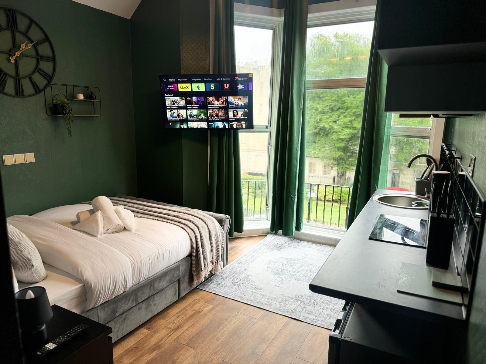 A hotel room with a bed, a television mounted on the wall, a window with curtains, and a small kitchen area.