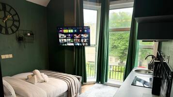 A hotel room with a bed, a television mounted on the wall, a window with curtains, and a small kitchen area.