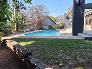 Une piscine entourée d’une pelouse, avec un mur en pierre et une terrasse en bois équipée de chaises longues.