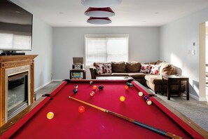 Een kamer met een pooltafel, een bank, een open haard en een raam met jaloezieën.