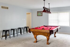Een pooltafel met krukken, een plafondventilator en een raam met jaloezieën.