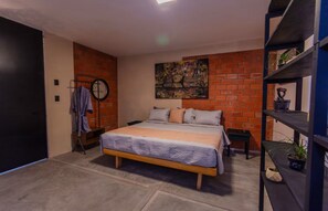 A bedroom with a bed, a brick wall, a painting, and a shelf with plants. - Loft with a King-size bed in San Felipe (Oaxaca de Juárez)