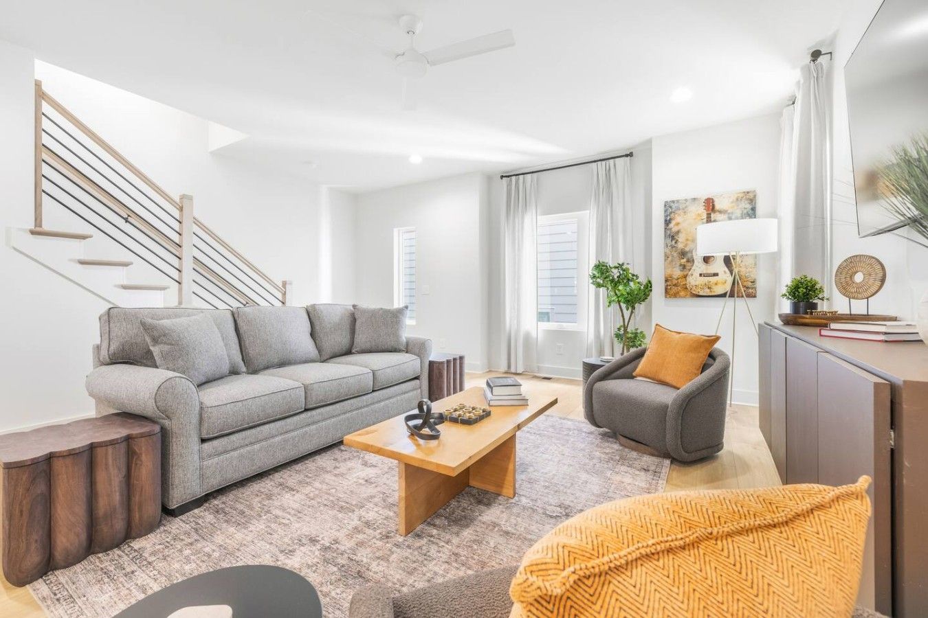 A modern living room with a grey sofa, a wooden coffee table, and a grey armchair. There is a staircase with wooden railings and a white ceiling fan.