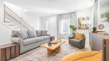 A modern living room with a grey sofa, a wooden coffee table, and a grey armchair. There is a staircase with wooden railings and a white ceiling fan.