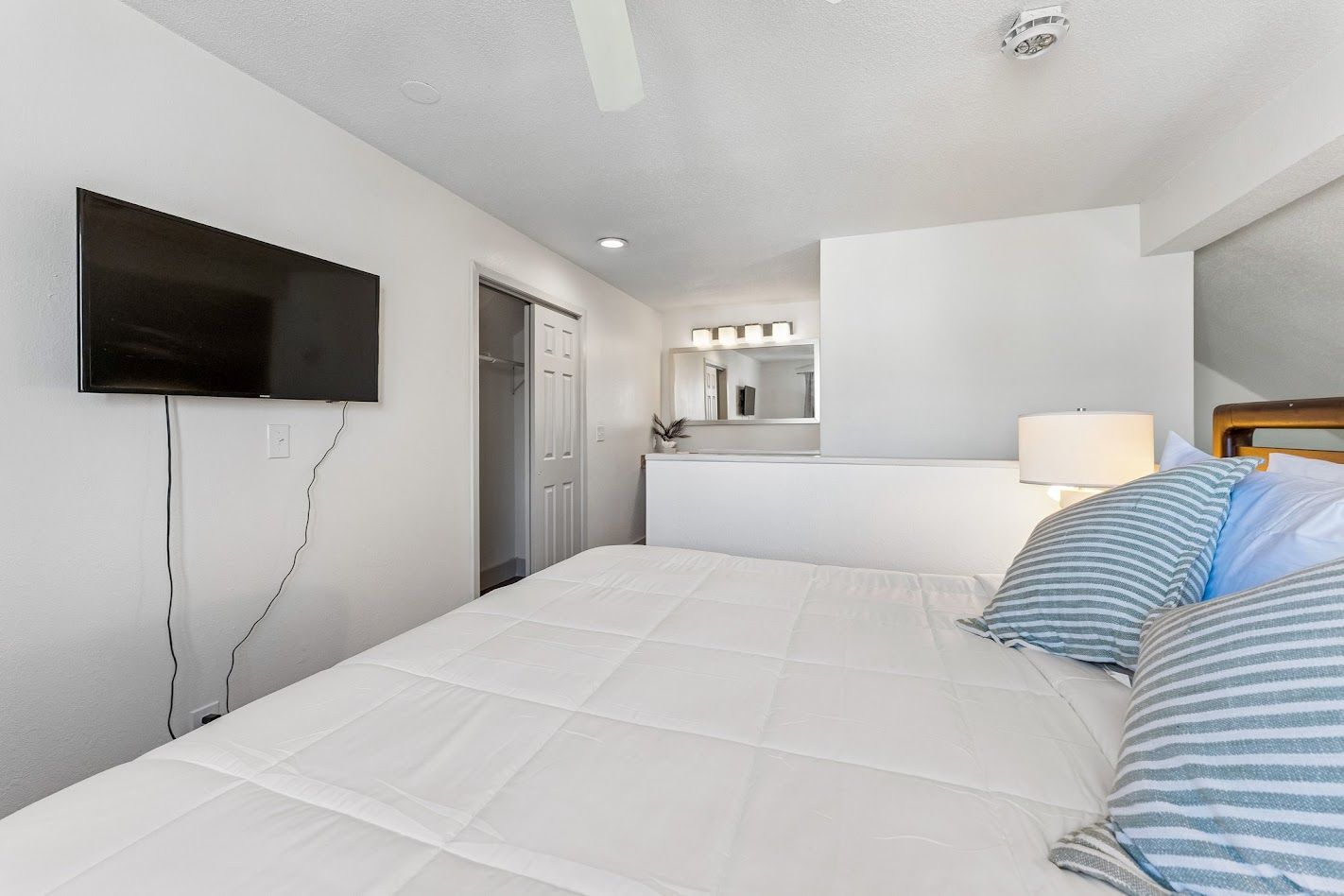 A hotel room with a bed, a TV mounted on the wall, a mirror above the bed, and a door leading to a bathroom.