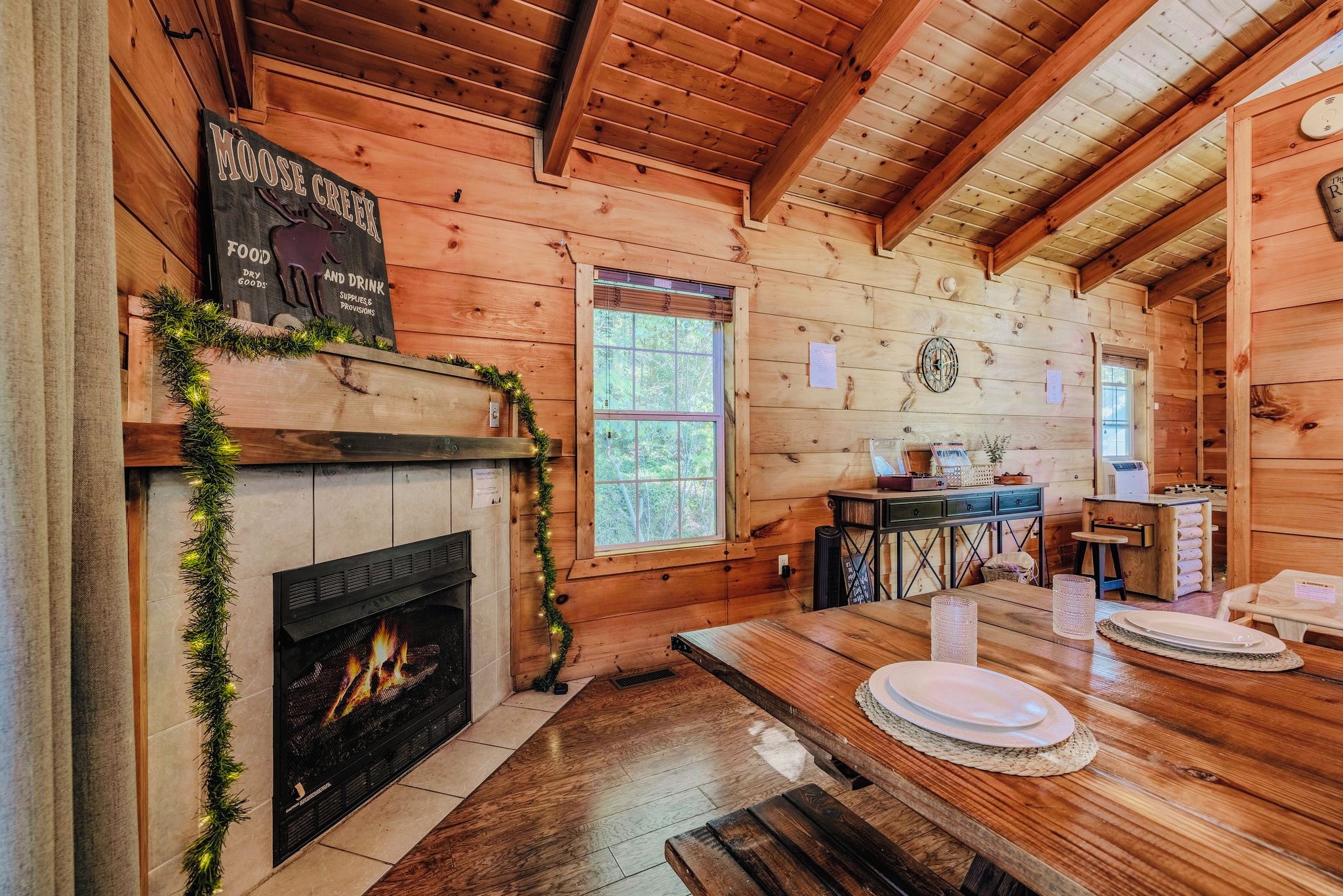 L’intérieur d’un chalet accueillant, avec une cheminée, des murs en bois, une table à manger dressée pour deux personnes et une fenêtre donnant sur la forêt.